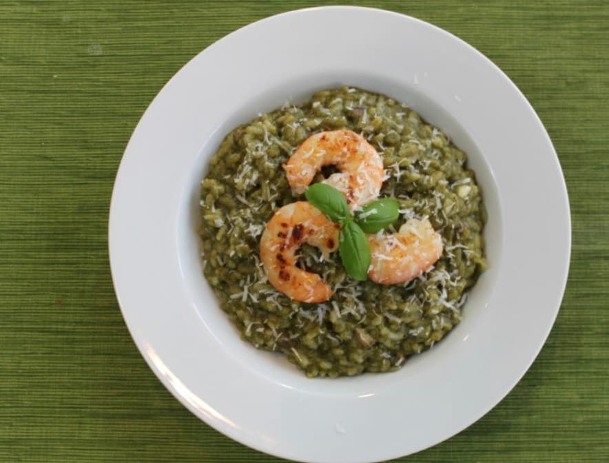 A plate of green pesto risotto topped with grilled shrimp, grated cheese, and a basil leaf, presented on a white plate over a green table mat