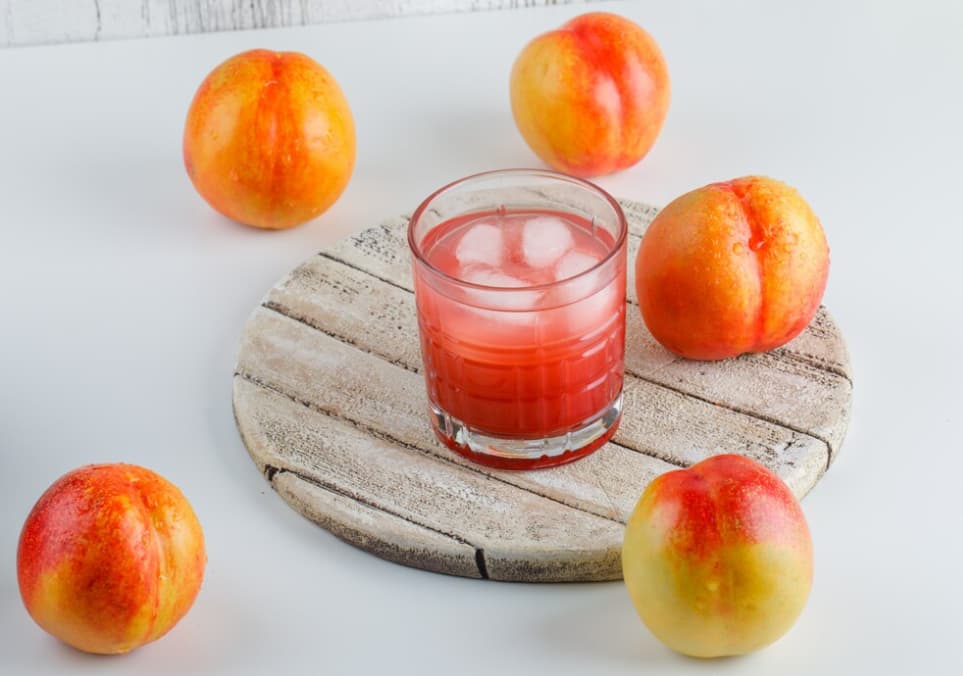 A glass of peach juice with ice on a wooden board surrounded by fresh peaches