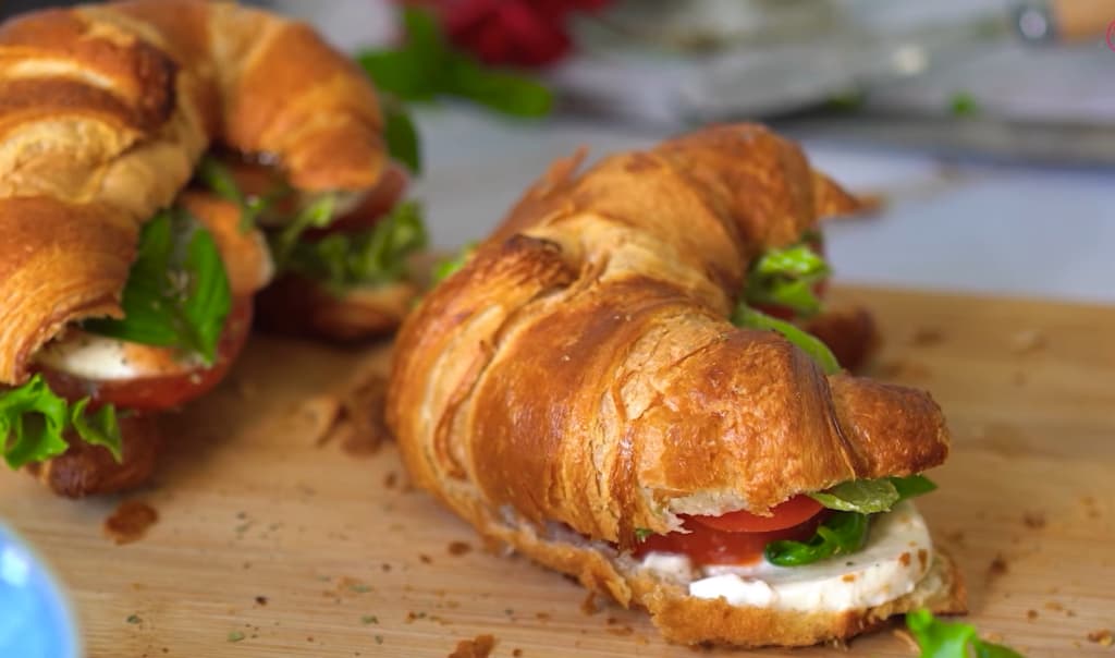 Close-up of a croissant sandwich with lettuce, tomato, and creamy cheese on a cutting board