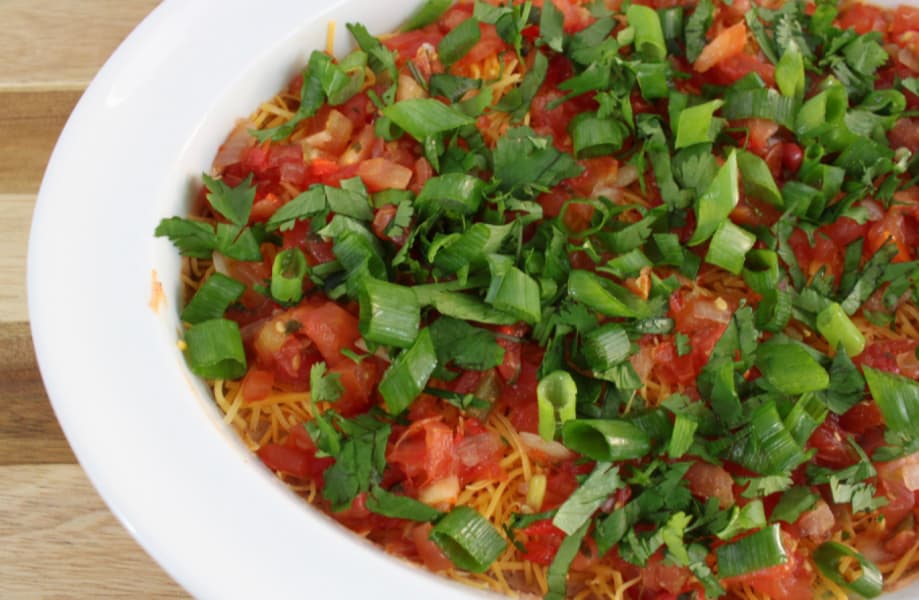 A Mexican layered dip in a white dish, topped with chopped tomatoes, green onions, and cilantro, ready to be served