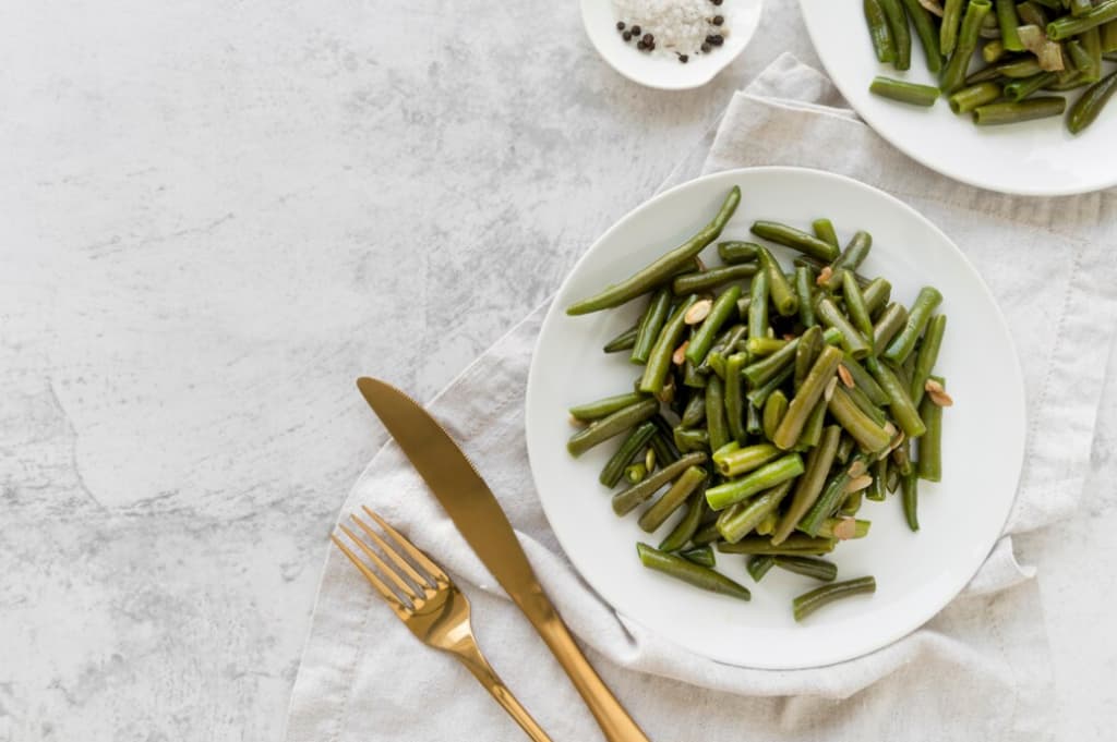 Green beans with pine nuts on a white plate, gold cutlery on a marble background
