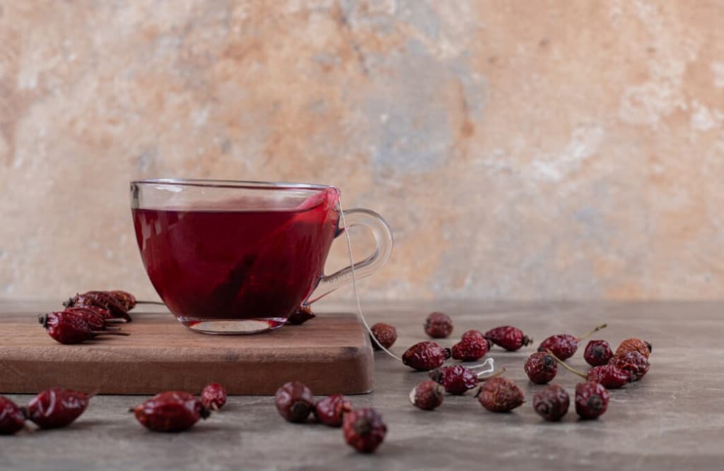 A clear glass cup of red tea on a wooden board, surrounded by dried rosehips