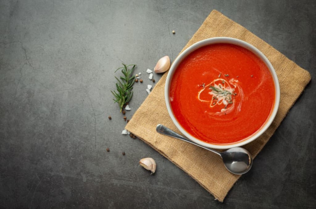 Bowl of creamy tomato soup with rosemary, garlic, and a spoon on a dark stone surface