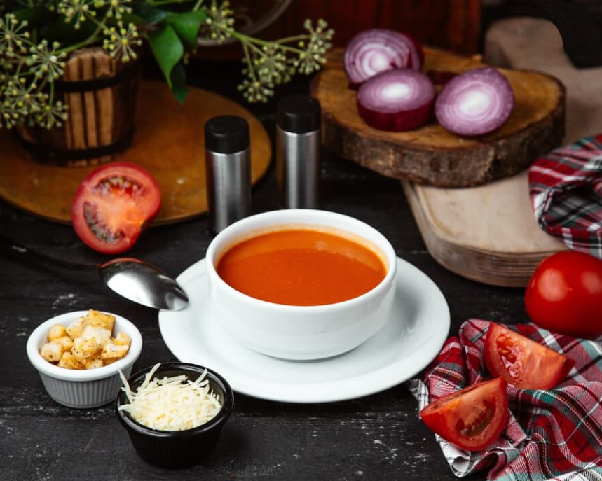 Tomato soup in a white bowl with croutons, cheese, and seasonings on a rustic table