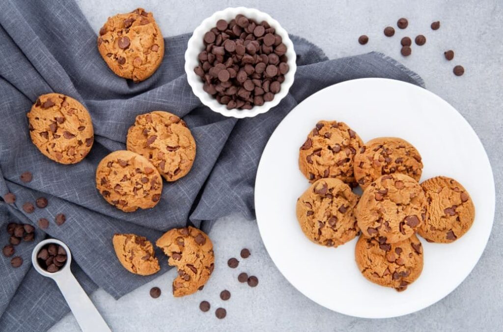 Chocolate chip cookies on a white plate with a bowl of chocolate chips on a grey cloth